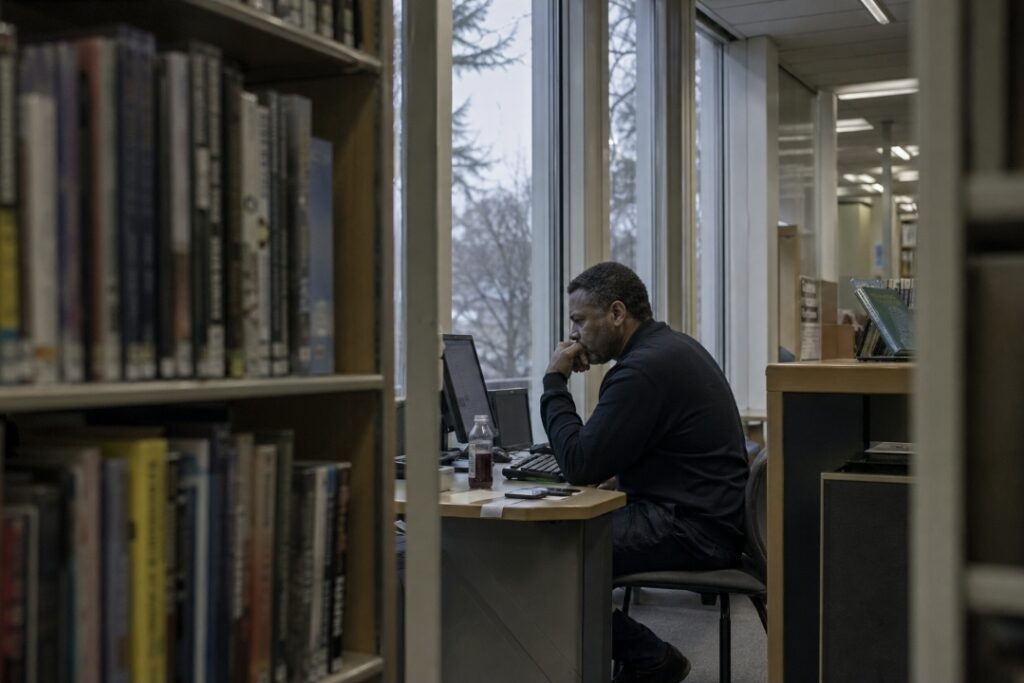 Man sitting at a public library computer in grey afternoon light with a dark juice bottle beside him, looking flat and irritable during a juice fast