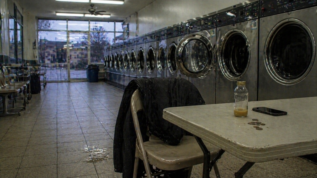 Laundrette table with an unfinished amber juice bottle, phone, coins, and coat on a chair under harsh fluorescent light