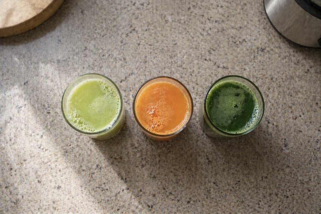 Three small glasses of pale green, orange, and dark green juice on a kitchen counter in natural daylight